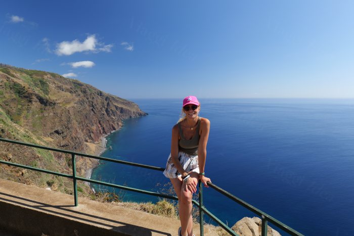 Donna con cappello rosa appoggiata a una ringhiera davanti a una costa alta e a mare turchese