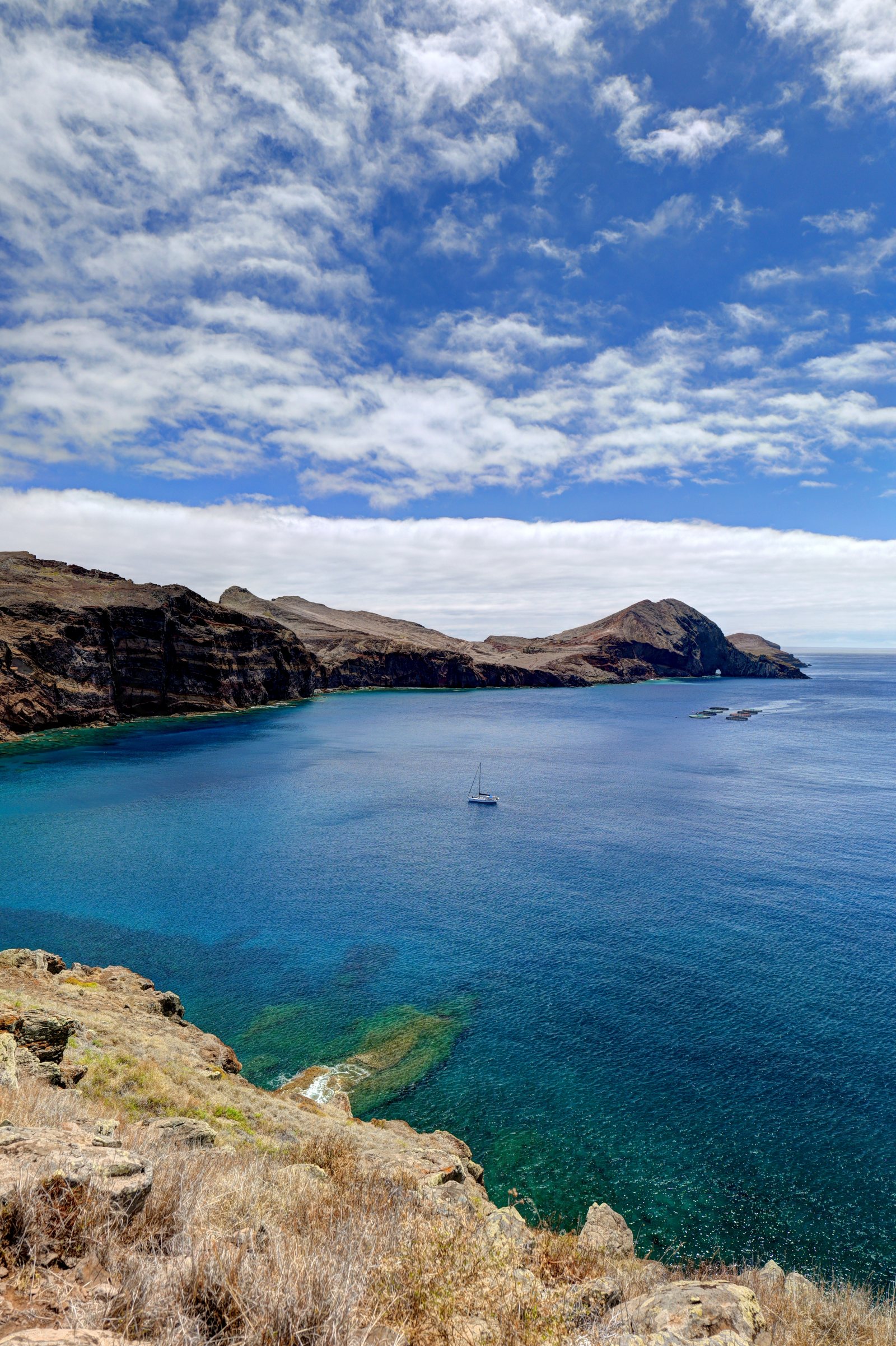 Baia con mare turchese, una barca a vela e scogliere rossastre sullo sfondo
